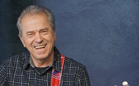 Ein älterer Mann spielt lächelnd Akustikgitarre vor einem blauen Hintergrund.