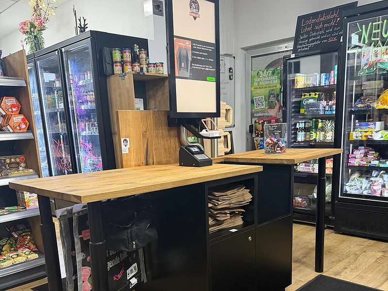 Innenansicht vom Genussmarkt 24/7 in Monheim - Regional und Gut Selbstbedienungskasse mit heller Holzplatte und digitalem Display in einem kleinen 24/7 Selbstbedienungs-Supermarkt mit Regalen und Kühlschränken gefüllt mit Ware im Hintergrund.