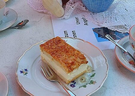 Gedeckter Tisch mit einem Stück Bienenstich-Torte auf dem Teller und einer Gabel im Vordergrund. Außerdem am Tisch in der Mitte des Bildes eine Porzellankanne mit Blumen, daneben eine Puppe mit rosa Schal, gefülltes Wasserglas, umgeben von weiteren Tellern mit Torten. Im Hintergrund der Oberkörper einer Person zu sehen.