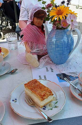 Gedeckter Tisch mit einem Stück Bienenstich-Torte auf dem Teller und einer Gabel im Vordergrund. Außerdem am Tisch in der Mitte des Bildes eine Porzellankanne mit Blumen, daneben eine Puppe mit rosa Schal, gefülltes Wasserglas, umgeben von weiteren Tellern mit Torten. Im Hintergrund der Oberkörper einer Person zu sehen.