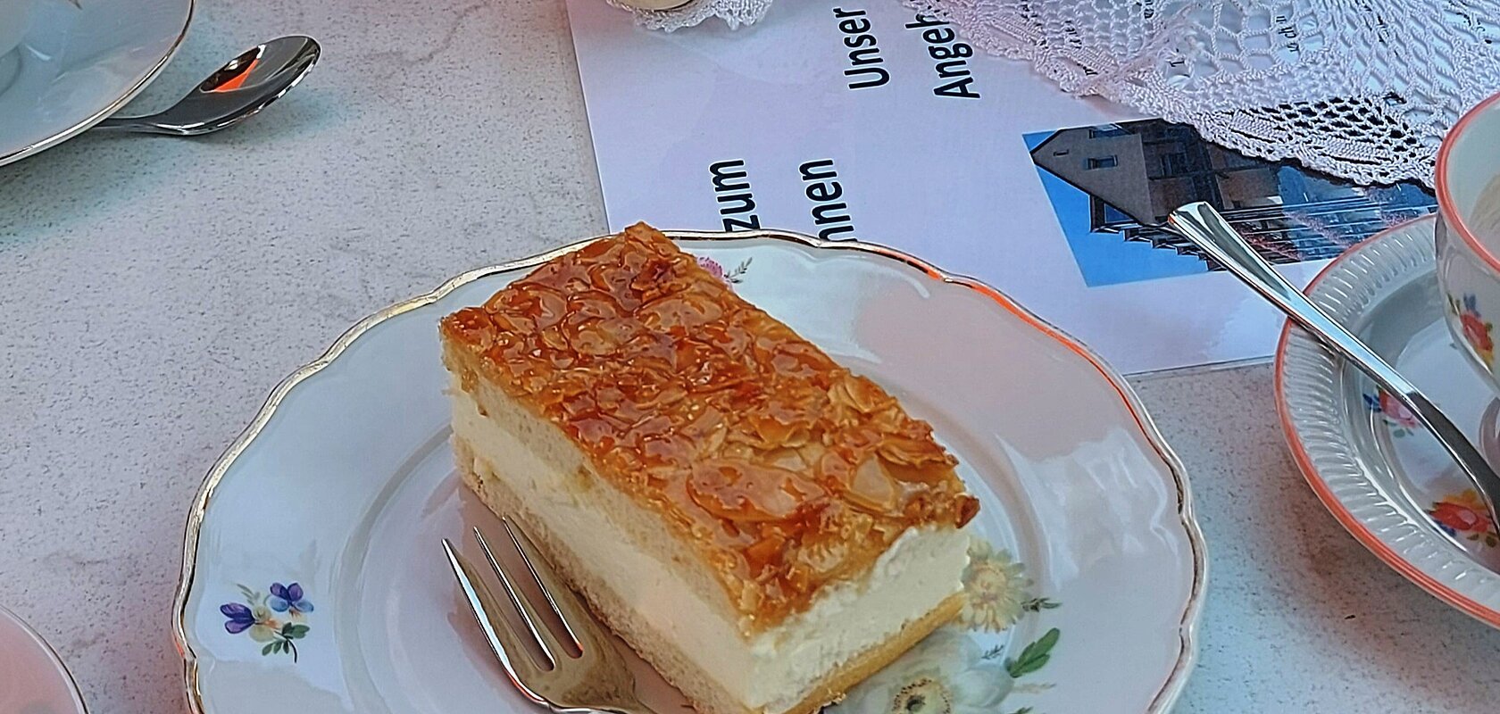 Gedeckter Tisch mit einem Stück Bienenstich-Torte auf dem Teller und einer Gabel im Vordergrund. Außerdem am Tisch in der Mitte des Bildes eine Porzellankanne mit Blumen, daneben eine Puppe mit rosa Schal, gefülltes Wasserglas, umgeben von weiteren Tellern mit Torten. Im Hintergrund der Oberkörper einer Person zu sehen.