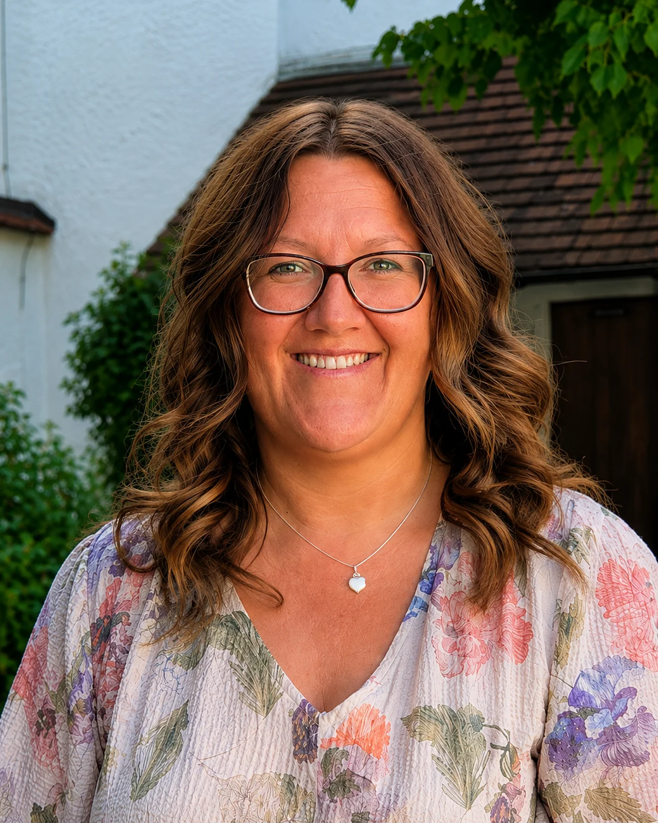 Jugendbeauftragte Kathrin Böswald Porträt einer lächelnden Frau mit Brille, braunen lockigen Haaren und floralem Oberteil vor Hausfassade und Grün. Erste Jugendbeauftragte der Stadt Monheim: Kathrin Böswald