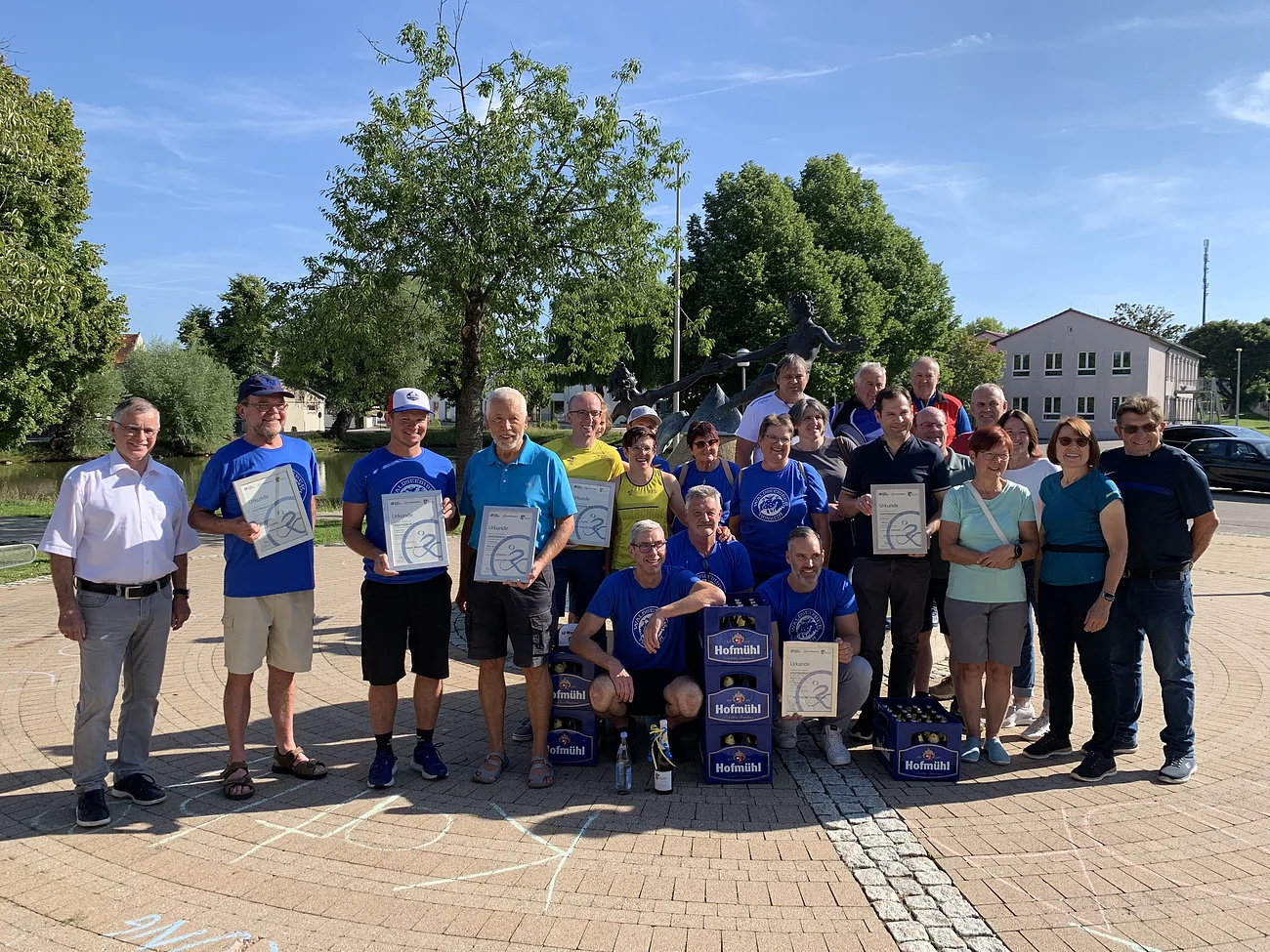 Gruppenfoto von 22 Personen im Freien auf einem gepflasterten Platz bei Sonnenschein, einige halten Urkunden, vor Bäumen und Gebäuden. Im Hintergrund sind rechts geparkte Fahrzeuge und links ein kleiner Weiher zu sehen.