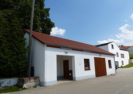 Weißes längliches Gebäude mit rotem Dach und WC-Schild über dem Eingang. Rechts vom Eingang ein Fenster, ein Holztor und weiteres Fenster daneben. Links vom Gebäude sind Bäume, vorderhalb ein gepflasterter Platz,  rechts im Bild ein Wohnhaus. Im Hintergrund strahlend blauer Himmel.