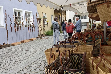 Marktstand in einer Innenstadt mit geflochtenen Körben, im Hintergrund Menschen und dekorative Metallstäbe vor bunten Häuserwänden.
