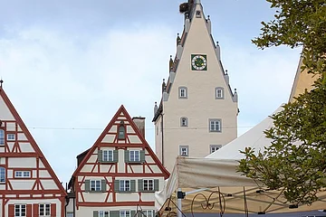 Markt mit Ständen und Besuchern vor Fachwerkhäusern und einem Turm mit Uhr und Storchnest.