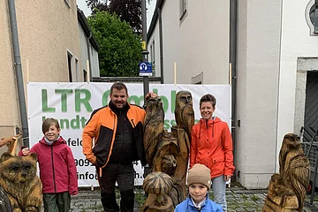 Vier Personen stehen hinter und neben geschnitzten Holzfiguren von Tieren auf einem gepflasterten Platz und blicken in die Kamera. Im Hintergrund zwei Wohnhäuser und eine kleine Gasse zu sehen, sowie ein Werbebanner an einem Bauzaun.