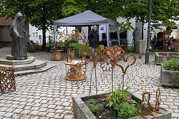 Marktstand mit Metallkunstwerken auf dem Pflasterplatz verteilt, Person am Pavillon mit schwarzem Dach, Marienstatue links, Bäume und Wohnhäuser im Hintergrund.