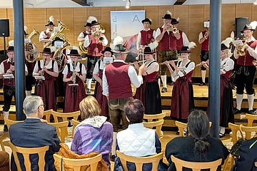 Blaskapelle in rot/schwarz/weißer Tracht spielt auf der Bühne, mit einer Holzverkleidung im Hintergrund, davor sitzen sechs Zuschauer auf Holzstühlen.