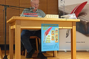 Älterer Mann liest an einem Holztisch mit Mikrofon, Plakat des Autorenclubs Donau-Ries hängt am Tisch. Neben dem Tisch ein großer Aufsteller des Autorenclubs Donau-Ries.