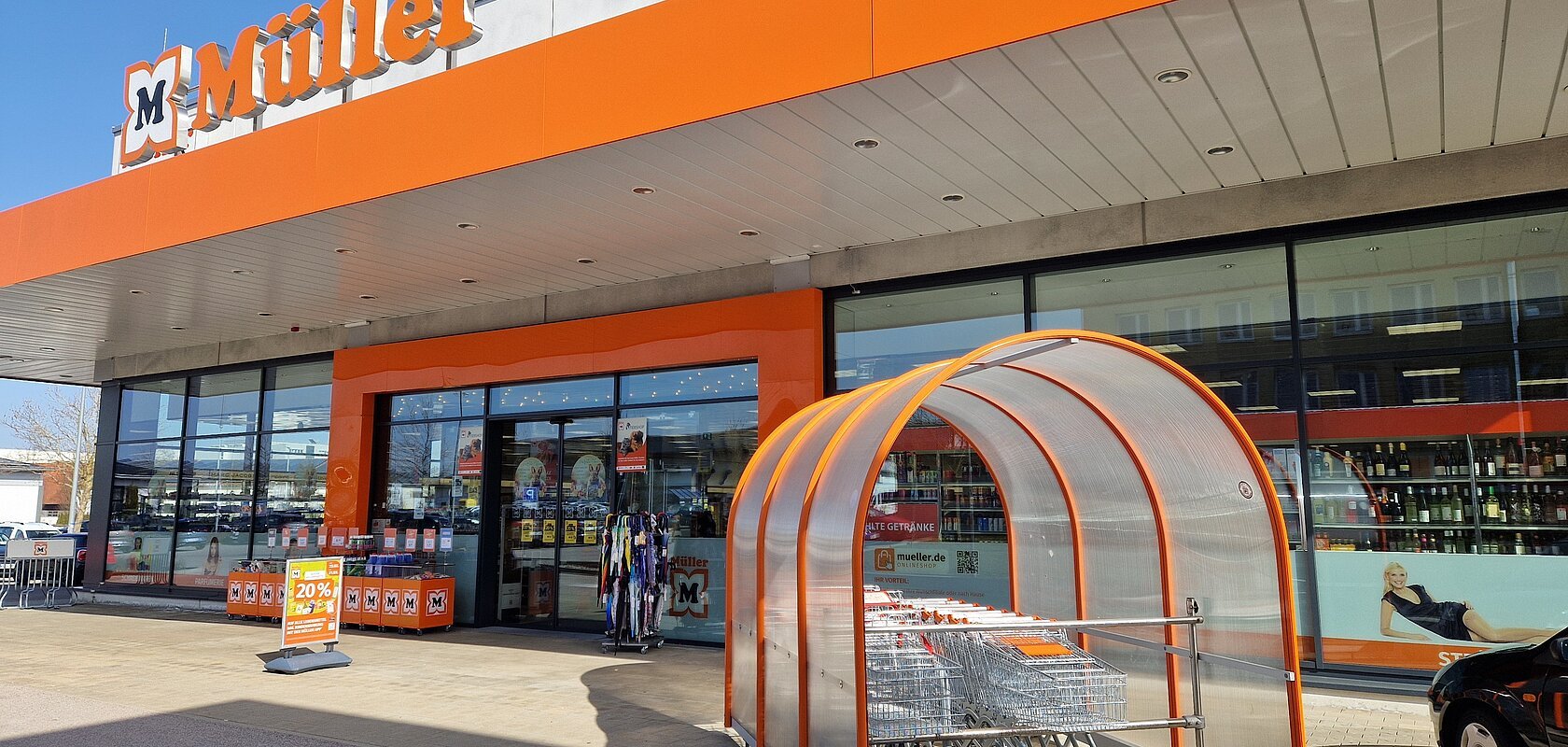 Eingangsbereich eines Müller-Marktes mit Einkaufswagen und orangen Markenzeichen "Müller" bei Sonnenschein.