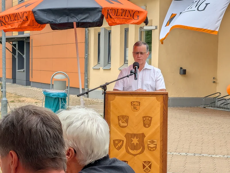 Mann in weißem Hemd mit Brille hält Rede am Rednerpult aus Holz mit 9 verschiedenen Wappen an der Front unter einem schwarz-orangem Sonnenschirm auf einem gepflastertem Platz. Davor zwei Personen mit schwarzen Kolping Monheim Shirts.