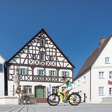 Der Marktplatz in Monheim: ein Fachwerkhaus zwischen zwei Gebäuden, ein Fahrradfahrer im Vordergrund fährt auf dem gepflastertem Platz, zwei Personen sitzen auf dem Rand eines Brunnens vor dem Fachwerkhaus. Auf dem linken Gebäude steht die Aufschrift "Stadtapotheke", auf dem rechten Gebäude die Aufschrift "Sparkasse".