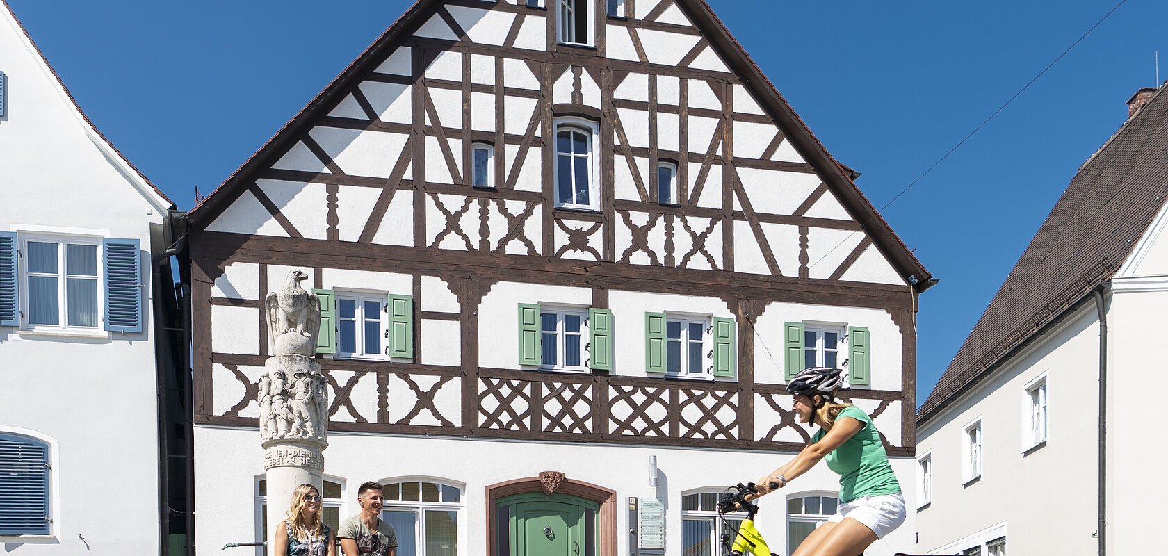 Der Marktplatz in Monheim: ein Fachwerkhaus zwischen zwei Gebäuden, ein Fahrradfahrer im Vordergrund fährt auf dem gepflastertem Platz, zwei Personen sitzen auf dem Rand eines Brunnens vor dem Fachwerkhaus. Auf dem linken Gebäude steht die Aufschrift "Stadtapotheke", auf dem rechten Gebäude die Aufschrift "Sparkasse".