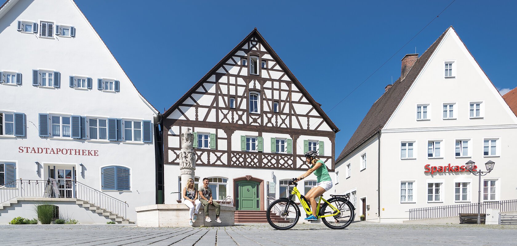 Der Marktplatz in Monheim: ein Fachwerkhaus zwischen zwei Gebäuden, ein Fahrradfahrer im Vordergrund fährt auf dem gepflastertem Platz, zwei Personen sitzen auf dem Rand eines Brunnens vor dem Fachwerkhaus. Auf dem linken Gebäude steht die Aufschrift "Stadtapotheke", auf dem rechten Gebäude die Aufschrift "Sparkasse".