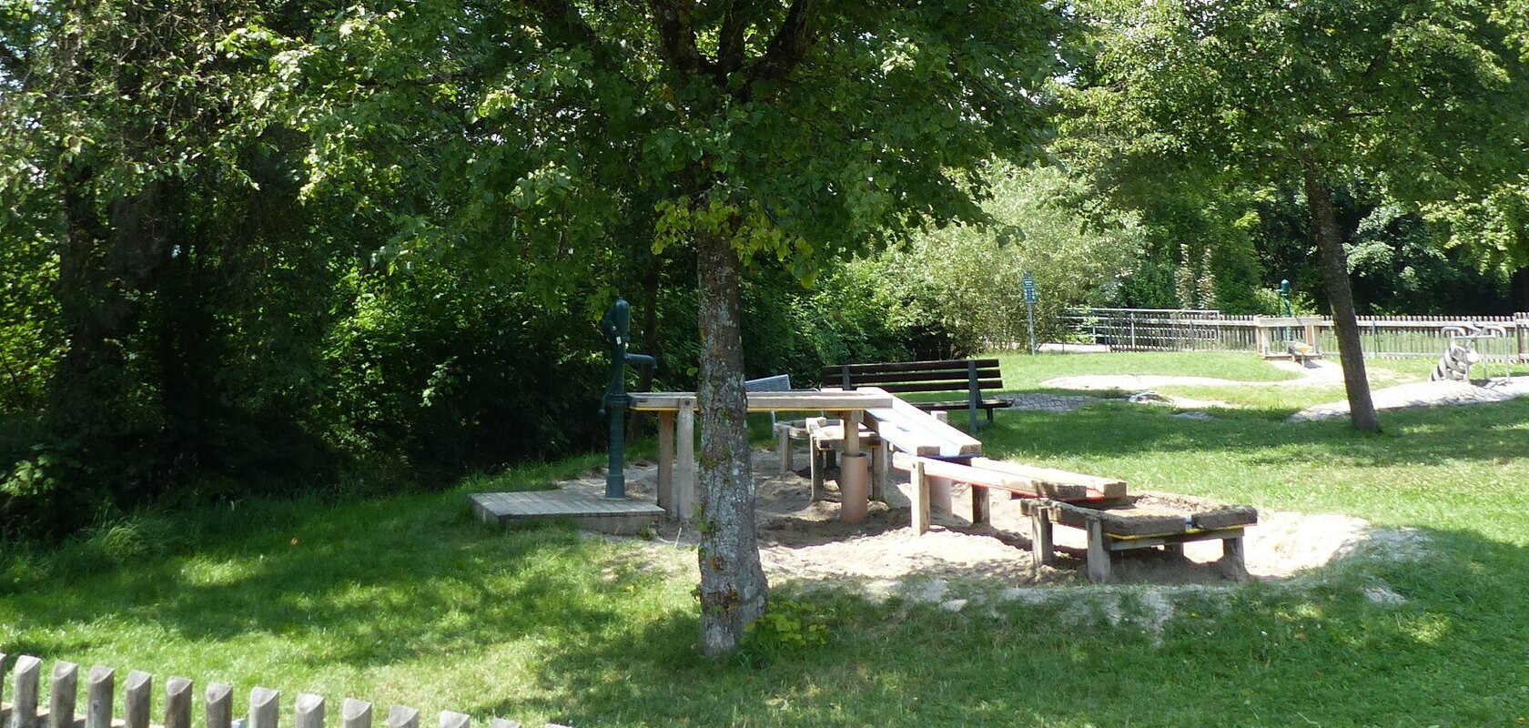 Wasserspielplatz mit Sand, Holztischen und Bänken, umgeben von Bäumen und einem Holzzaun. Informationstafel links.