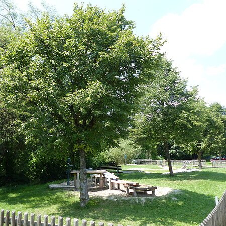 Wasserspielplatz mit Sand, Holztischen und Bänken, umgeben von Bäumen und einem Holzzaun. Informationstafel links.