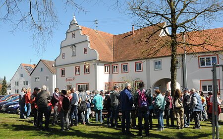 Gruppe von Menschen steht auf einer Wiese vor einem historischen Gebäude mit rotem Ziegeldach bei klarem Himmel.Senioren wandern mit SoMit Verein am 08.04.2026 in Fünfstetten