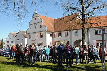 Gruppe von Menschen steht auf einer Wiese vor einem historischen Gebäude mit rotem Ziegeldach bei klarem Himmel.Senioren wandern mit SoMit Verein am 08.04.2026 in Fünfstetten