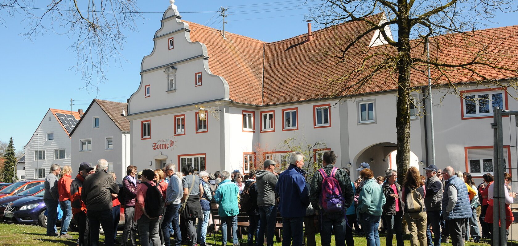 Gruppe von Menschen steht auf einer Wiese vor einem historischen Gebäude mit rotem Ziegeldach bei klarem Himmel.Senioren wandern mit SoMit Verein am 08.04.2026 in Fünfstetten