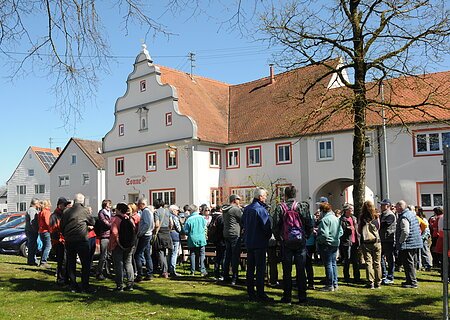 Gruppe von Menschen steht auf einer Wiese vor einem historischen Gebäude mit rotem Ziegeldach bei klarem Himmel.Senioren wandern mit SoMit Verein am 08.04.2026 in Fünfstetten