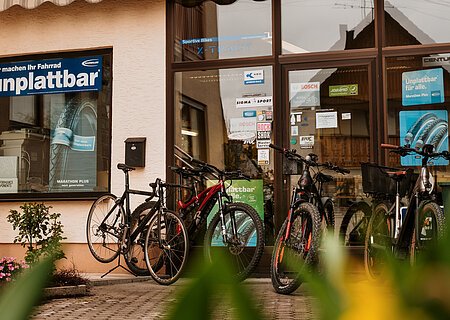 Fahrradgeschäft mit mehreren Fahrrädern vor dem Eingang und Werbeplakaten zu pannensicheren Reifen auf der Glasscheibe des Geschäfts. Im Vordergrund unscharf grüne Pflanzen zu sehen.