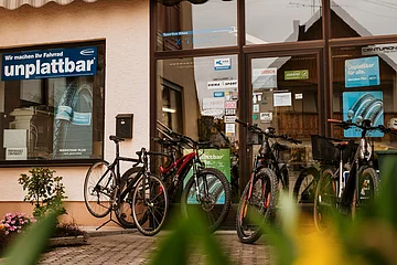 Fahrradgeschäft mit mehreren Fahrrädern vor dem Eingang und Werbeplakaten zu pannensicheren Reifen auf der Glasscheibe des Geschäfts. Im Vordergrund unscharf grüne Pflanzen zu sehen.