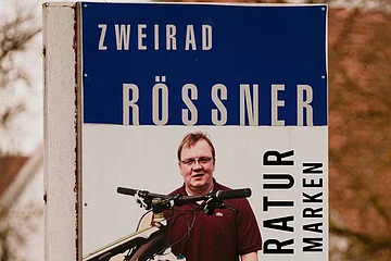 Werbeschild für Fahrradreparatur mit Mann und Fahrrad, Text „Zweirad Rössner Reparatur aller Marken“ und Telefonnummer.