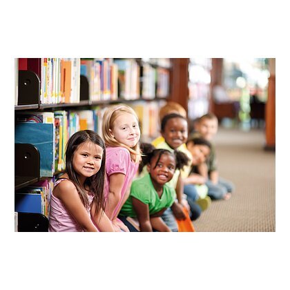 Kinder sitzen in einer Reihe auf dem Boden einer Bibliothek, Bücherregale im Hintergrund.