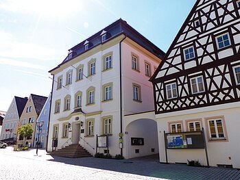 Straßenansicht der Monheimer Innenstadt: Im Vordergrund das Rathaus der Stadt Monheim, mit weiteren mehreren Gebäuden, darunter ein Fachwerkhaus, das man Schindlerhaus nennt.