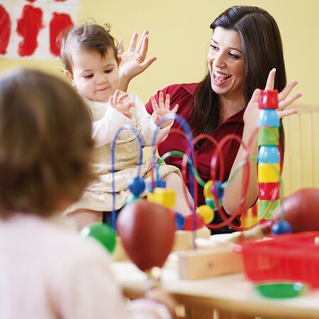 Eine Erwachsene Frau spielt am Tisch mit drei Kindern in einem Raum mit buntem Spielzeug. In der Mitte des Tisches steht ein Motorikwürfel und Bauklötze.