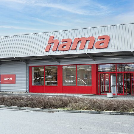 Graue Fassade eines Outlet-Geschäfts mit rotem Schriftzug "Hama", roten Eingangstüren und roten Fensterumrandungen, bei bewölktem Himmel. Vor dem Geschäft eine Hecke entlang einer Straße.