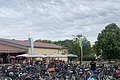 Parkplatz mit vielen Fahrrädern nebeneinander, im Hintergrund Menschen unter Sonnenschirmen vor einem gelben Gebäude, bewölkter Himmel.