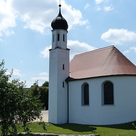 Weißes Kirchgebäude mit abgerundetem Grundriss und kleinem Turm mit schwarzer Haube bei blauem Himmel, umgeben von grünen Bäumen und wiese. Im Hintergrund weitere Häuser.