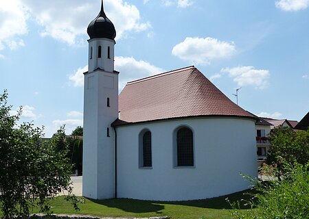 Weißes Kirchgebäude mit abgerundetem Grundriss und kleinem Turm mit schwarzer Haube bei blauem Himmel, umgeben von grünen Bäumen und wiese. Im Hintergrund weitere Häuser.