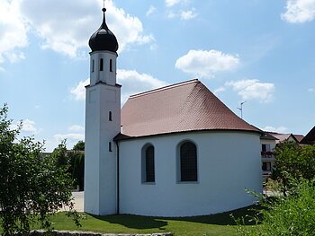 Weißes Kirchgebäude mit abgerundetem Grundriss und kleinem Turm mit schwarzer Haube bei blauem Himmel, umgeben von grünen Bäumen und wiese. Im Hintergrund weitere Häuser.