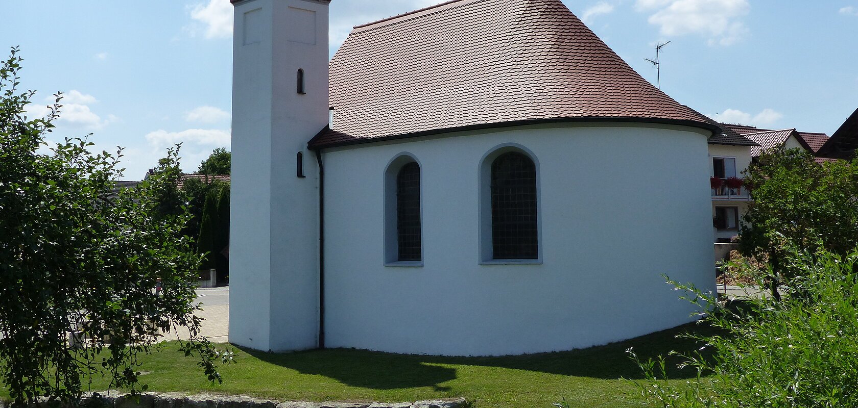 Weißes Kirchgebäude mit abgerundetem Grundriss und kleinem Turm mit schwarzer Haube bei blauem Himmel, umgeben von grünen Bäumen und wiese. Im Hintergrund weitere Häuser.
