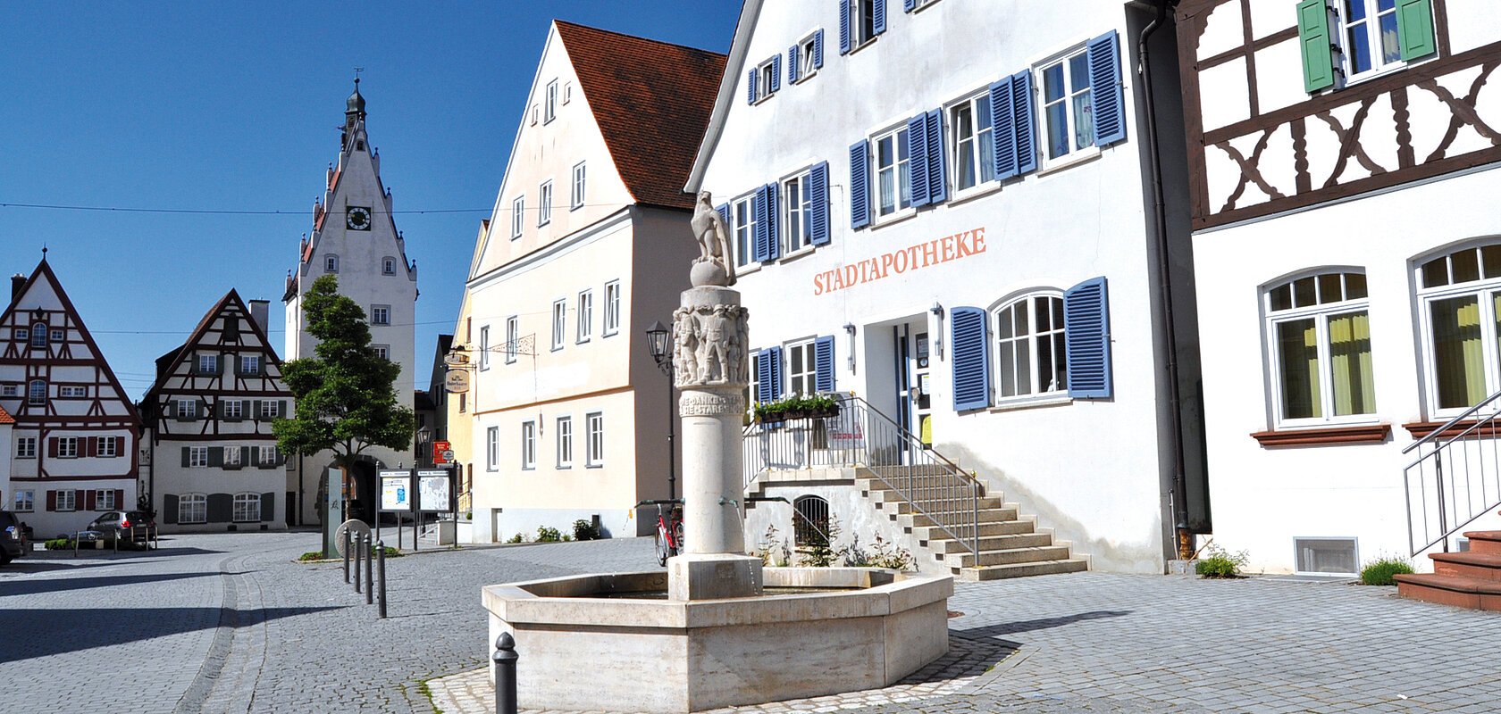 Historischer gepflasterter Marktplatz mit Fachwerkhäusern auf einem Gebäude steht die Aufschrift "Stadtapotheke". Ein Brunnen mit einer Steinskulptur steht davor. Links verläuft eine Straße.