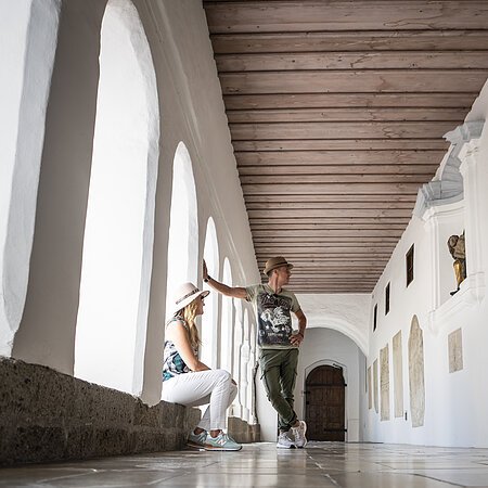 Zwei Personen mit Hüten in einem Arkadengang, dem Kreuzgang im ehemaligen Kloster in Monheim, mit weißen Bögen und Holzbalkendecke.