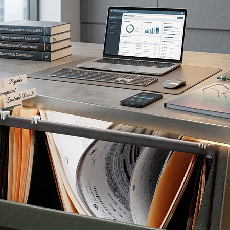 Bürotisch mit offenem Laptop, Tastatur, Maus, Smartphone, Brille und  einem Stempel. Im Vordergrund sind Aktenordner mit Beschriftungen zu Ämtern zu sehen.