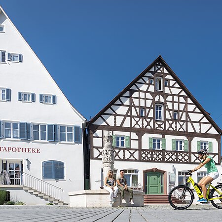 Der Marktplatz in Monheim: ein Fachwerkhaus zwischen zwei Gebäuden, ein Fahrradfahrer im Vordergrund fährt auf dem gepflastertem Platz, zwei Personen sitzen auf dem Rand eines Brunnens vor dem Fachwerkhaus. Auf dem linken Gebäude steht die Aufschrift "Stadtapotheke", auf dem rechten Gebäude die Aufschrift "Sparkasse".