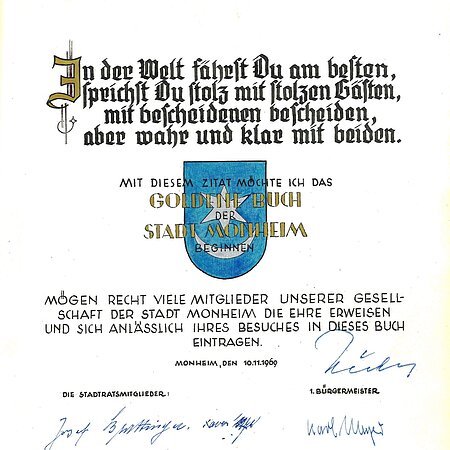 Erster Eintrag im Goldenen Buch der Stadt Monheim: Oben ein Zitat, darunter das Wappen der Stadt Monheim in blau. Darunter ein weiterer Text, datiert mit 10.11.1969, mit etlichen Unterschriften darunter.