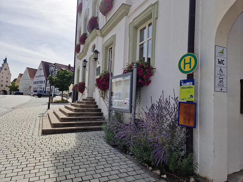 Im Vordergrund ein Haltestellenschild an weißem Gebäude, sowie ein weiteres blau-grünes Schild mit der Aufschrift SoMit-Bürgerbus. Darunter ein Beet mit Blumen, links davon eine Treppe auf gepflastertem Platz in kleinstädtischer Umgebung.