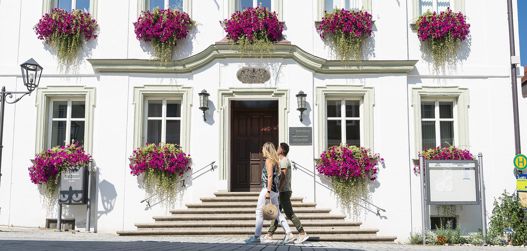 Das Rathaus in Monheim: Ein weißes dreistöckiges Gebäude mit mehreren Fenstern, an denen Blumenkästen mit pinken Blumen hängen. Zwei Personen gehen davor vorbei. Links und rechts davon jeweils ein weiteres Haus.