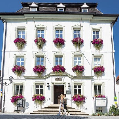 Das Rathaus in Monheim: Ein weißes dreistöckiges Gebäude mit mehreren Fenstern, an denen Blumenkästen mit pinken Blumen hängen. Zwei Personen gehen davor vorbei. Links und rechts davon jeweils ein weiteres Haus.