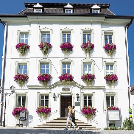 Das Rathaus in Monheim: Ein weißes dreistöckiges Gebäude mit mehreren Fenstern, an denen Blumenkästen mit pinken Blumen hängen. Zwei Personen gehen davor vorbei. Links und rechts davon jeweils ein weiteres Haus.