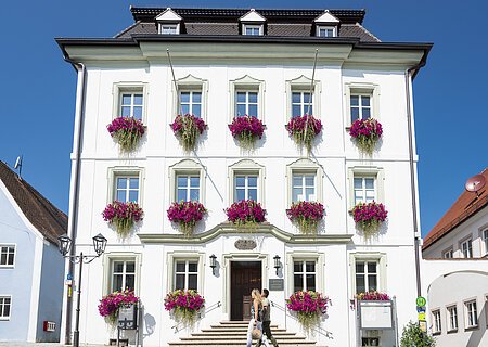 Das Rathaus in Monheim: Ein weißes dreistöckiges Gebäude mit mehreren Fenstern, an denen Blumenkästen mit pinken Blumen hängen. Zwei Personen gehen davor vorbei. Links und rechts davon jeweils ein weiteres Haus.