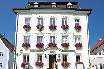 Das Rathaus in Monheim: Ein weißes dreistöckiges Gebäude mit mehreren Fenstern, an denen Blumenkästen mit pinken Blumen hängen. Zwei Personen gehen davor vorbei. Links und rechts davon jeweils ein weiteres Haus.