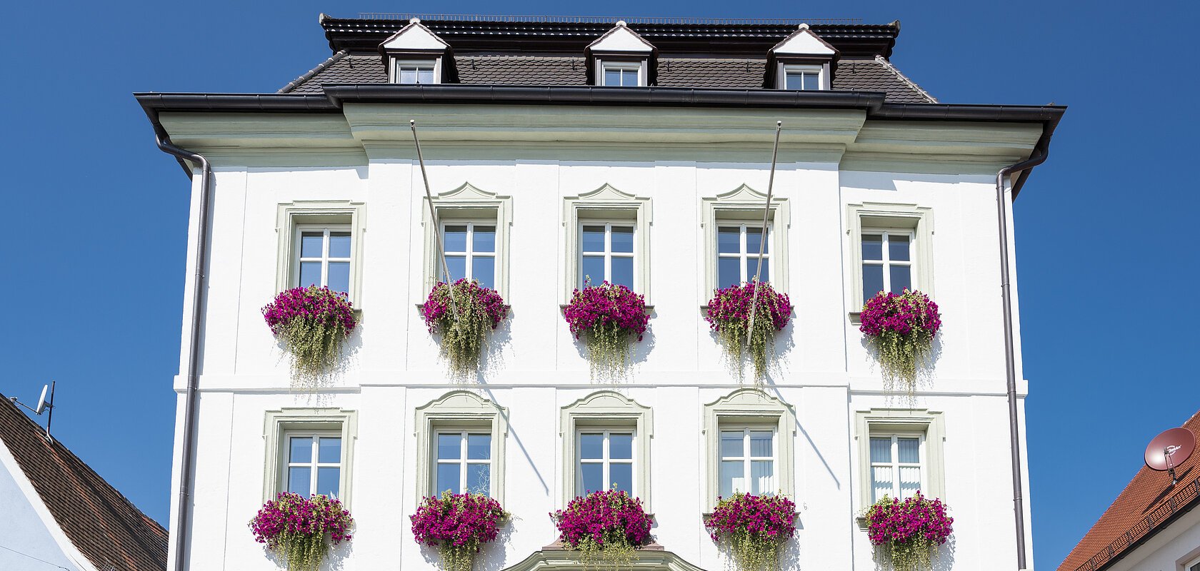 Das Rathaus in Monheim: Ein weißes dreistöckiges Gebäude mit mehreren Fenstern, an denen Blumenkästen mit pinken Blumen hängen. Zwei Personen gehen davor vorbei. Links und rechts davon jeweils ein weiteres Haus.