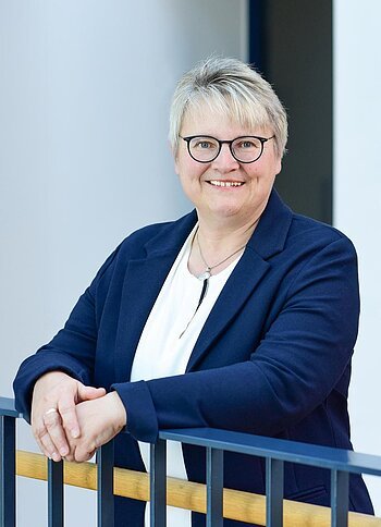 Porträt einer lächelnden Frau mit kurzen grauen Haaren, Brille, weißem Hemd und blauem Blazer, die an einem Geländer lehnt.
2. Bürgermeisterin der Stadt Monheim